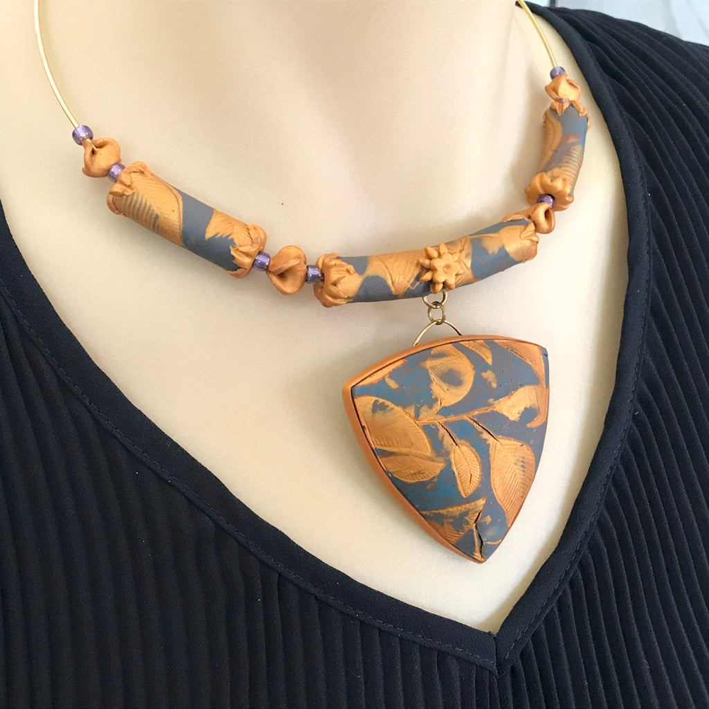 Handmade blue grey and gold leaf pendant necklace worn on neckline, featuring a lightweight textured botanical pendant with matching beaded collar.