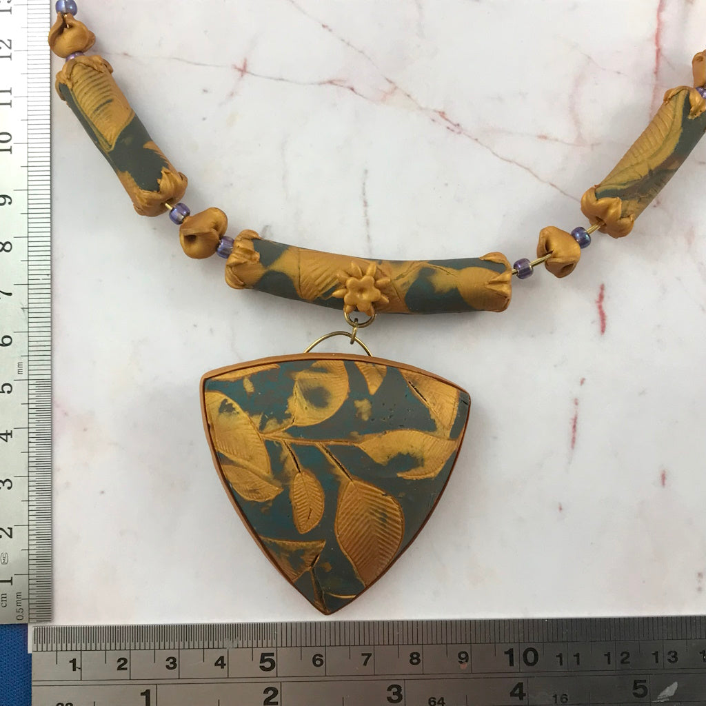 Handmade blue grey and gold leaf pendant necklace laid flat with ruler, featuring embossed botanical leaf design and matching beaded collar.