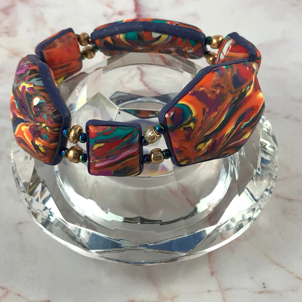 Artisan handmade polymer clay bracelet displayed on crystal stand with bold sunset swirl pattern and decorative metallic beads.