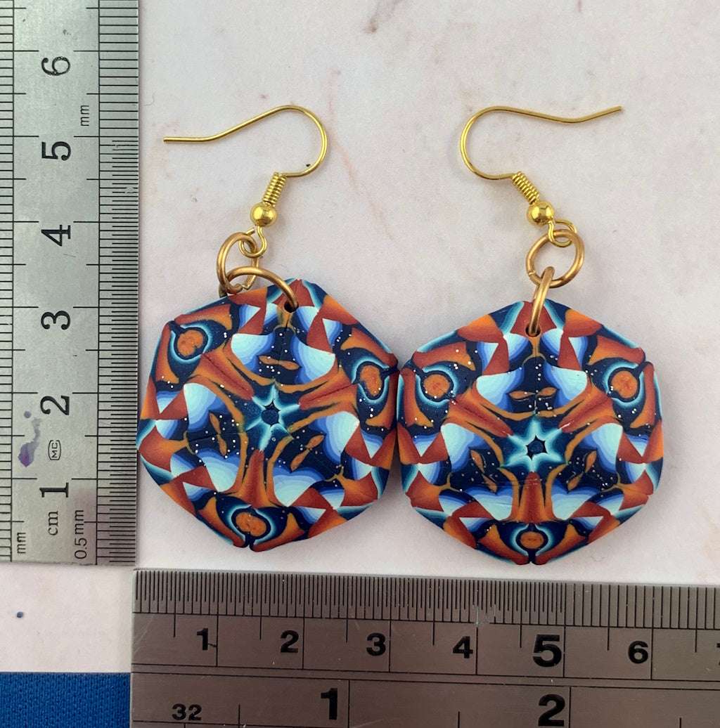 Handmade geometric polymer clay drop earrings with rust orange, teal and navy mandala pattern shown with ruler for size reference and gold hooks.