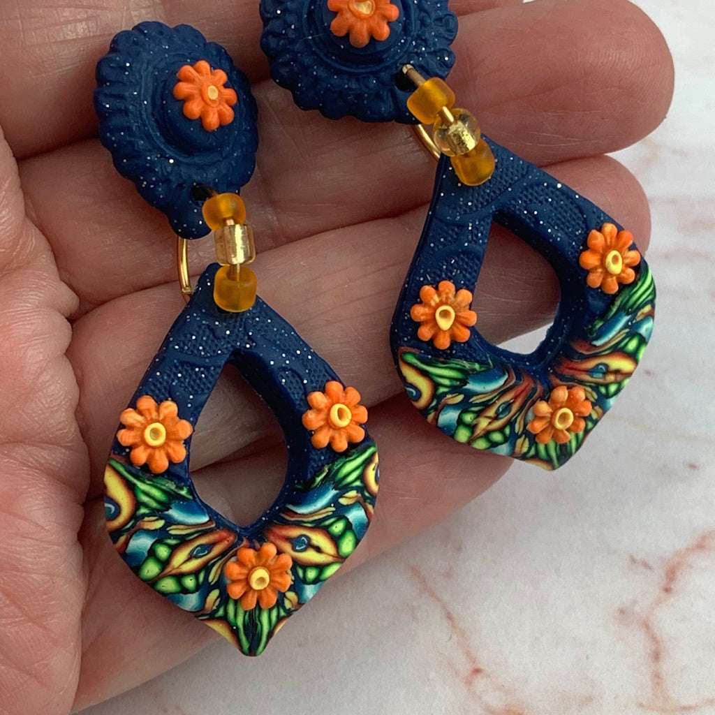 Close-up of award-winning Rennique hand-sculpted blue and orange floral drop earrings with glass bead accents in hand.