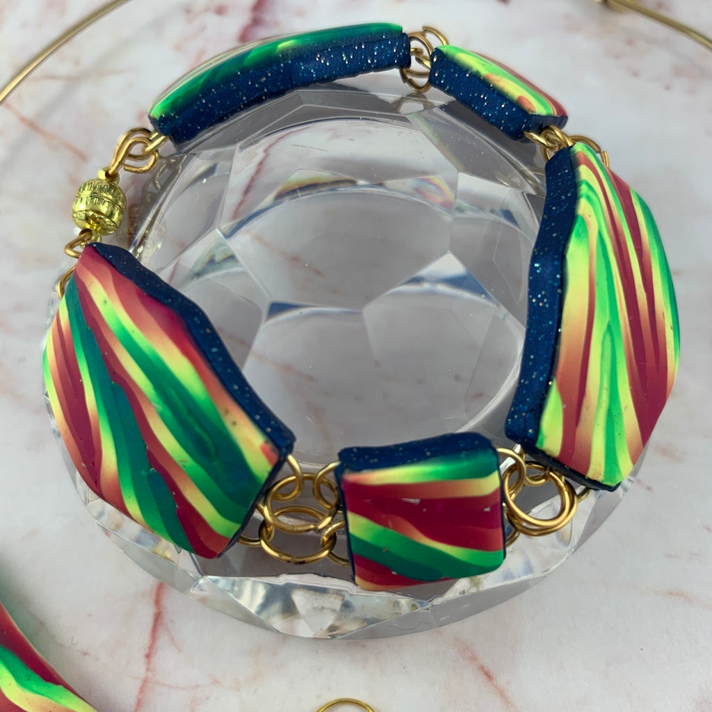 Close-up of Rennique handmade linked statement bracelet featuring bold striped polymer clay panels in vibrant green, pink and teal with gold hardware and textured navy edges.