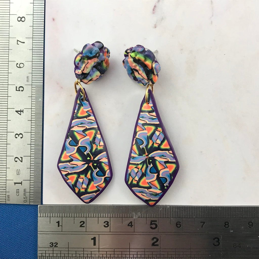 Rennique Studio one-of-a-kind long drop polymer clay earrings featuring kaleidoscope wearable art design in purple, pastel peach, blue, and neon rainbow tones, shown with ruler for size reference.