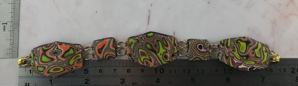 Handmade polymer clay bracelet laid flat with ruler, featuring ripple marbled pattern in coral, lime green, plum and black with gold wire links.