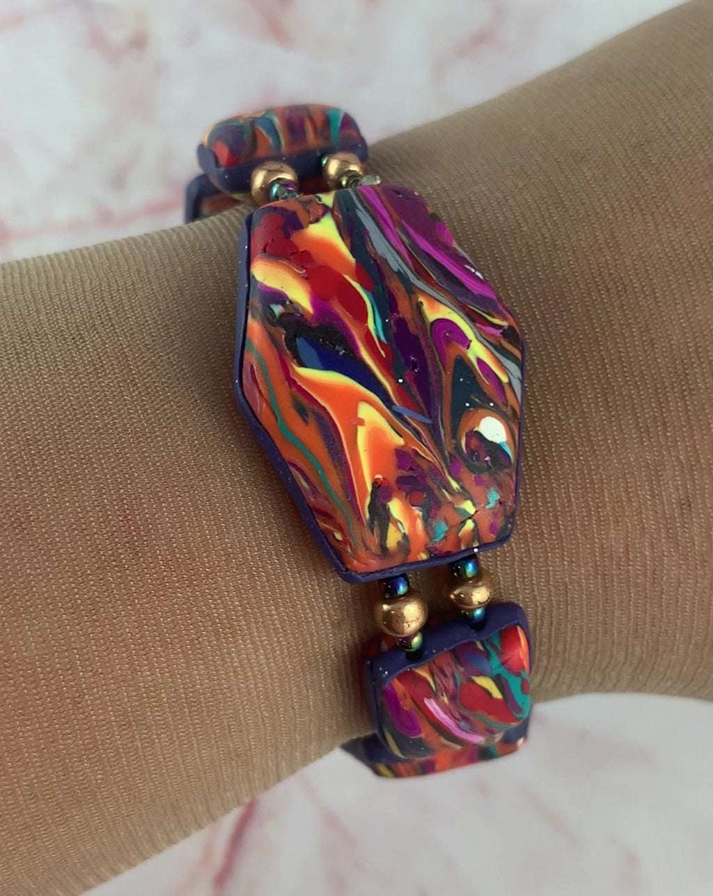 Handmade polymer clay statement bracelet worn on wrist featuring vibrant sunset swirl tones of orange, magenta, teal, and navy with metallic bead accents.